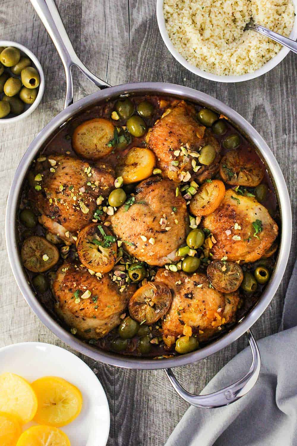 An overhead view of a large silver skillet filled with chicken tagine with preserved lemons and olives next to a bowl of couscous and a grey napkin.