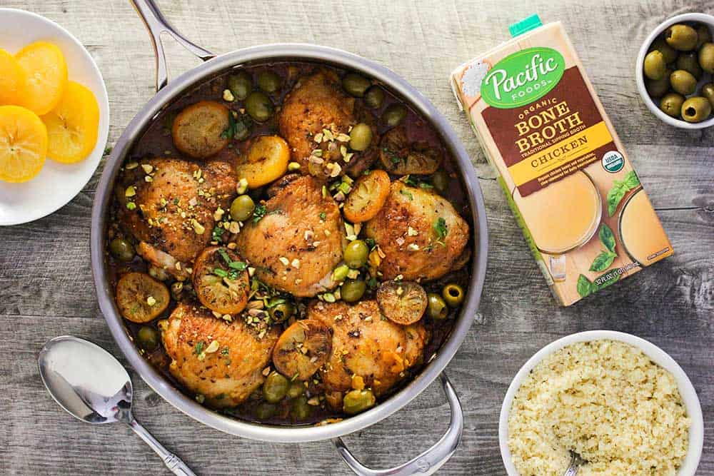 A large silver skillet filled with chicken tagine with preserved lemons and olives, next to a bowl of couscous and a box of Pacific Foods organic chicken broth. 