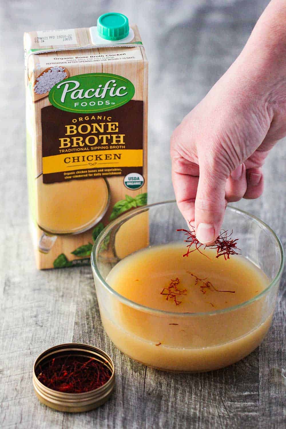 A hand sprinkling saffron threads into a glass bowl of chicken broth next to a box of Pacific Foods chicken broth. 