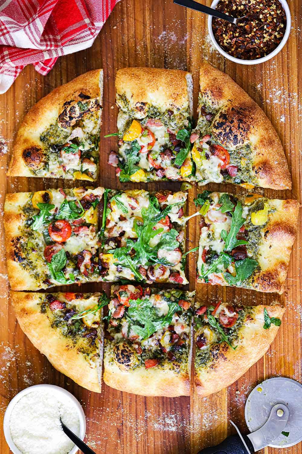 A overhead view of a BLT pizza cut into squares on a wooden cutting board with a small bowl of red pepper flakes and a bowl of Parmesan cheese nearby.