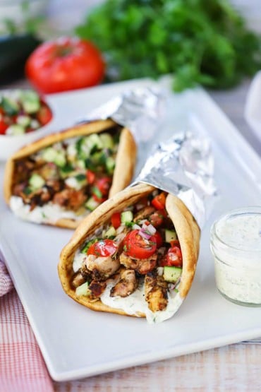 Two chicken gyros wrapped in foil on a white platter next to a small jar filled with tzatziki sauce with a spoon in, with a whole tomato and parsley in the background.