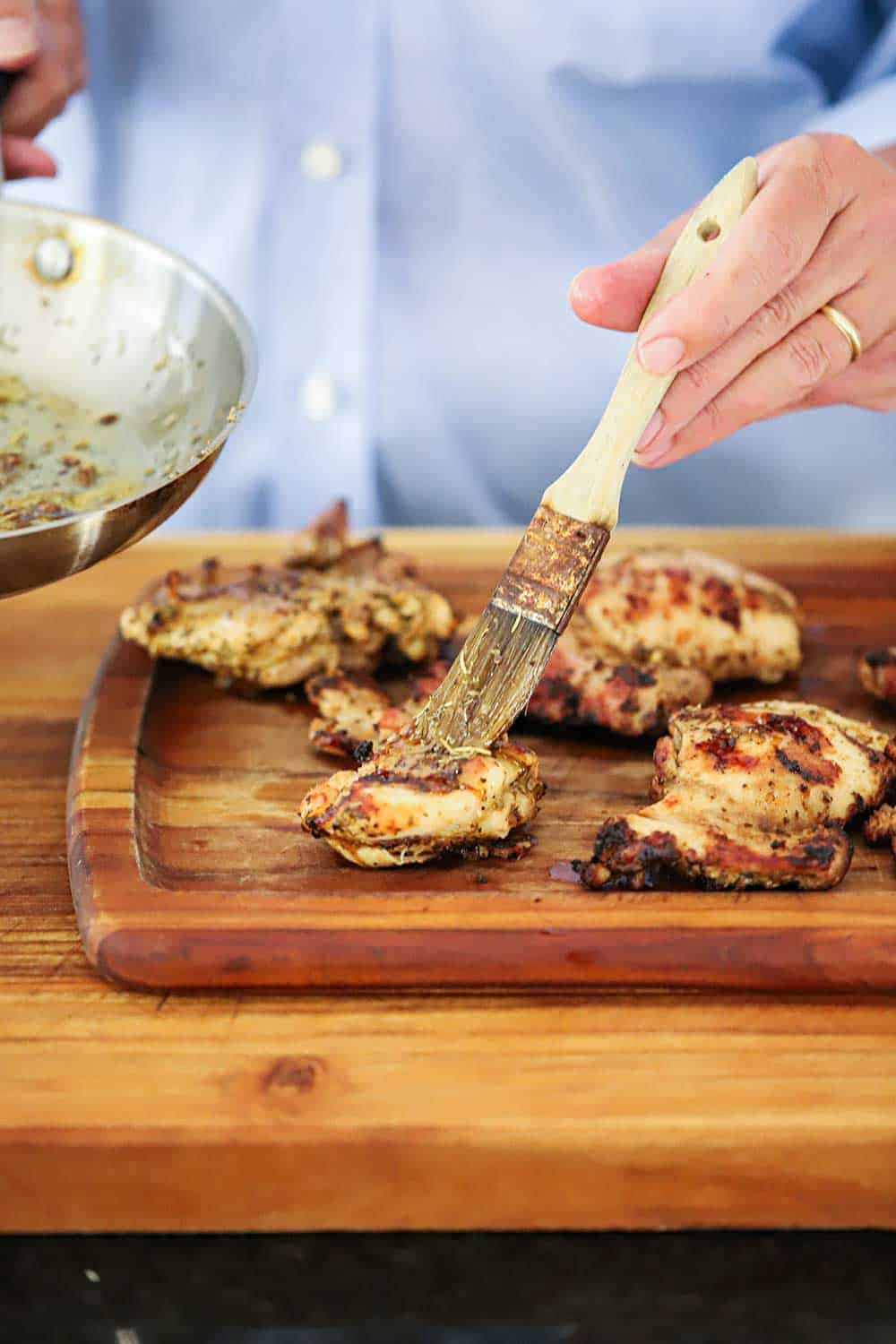 A hand brushing infused oil over grilled chicken thighs on a wooden cutting board.