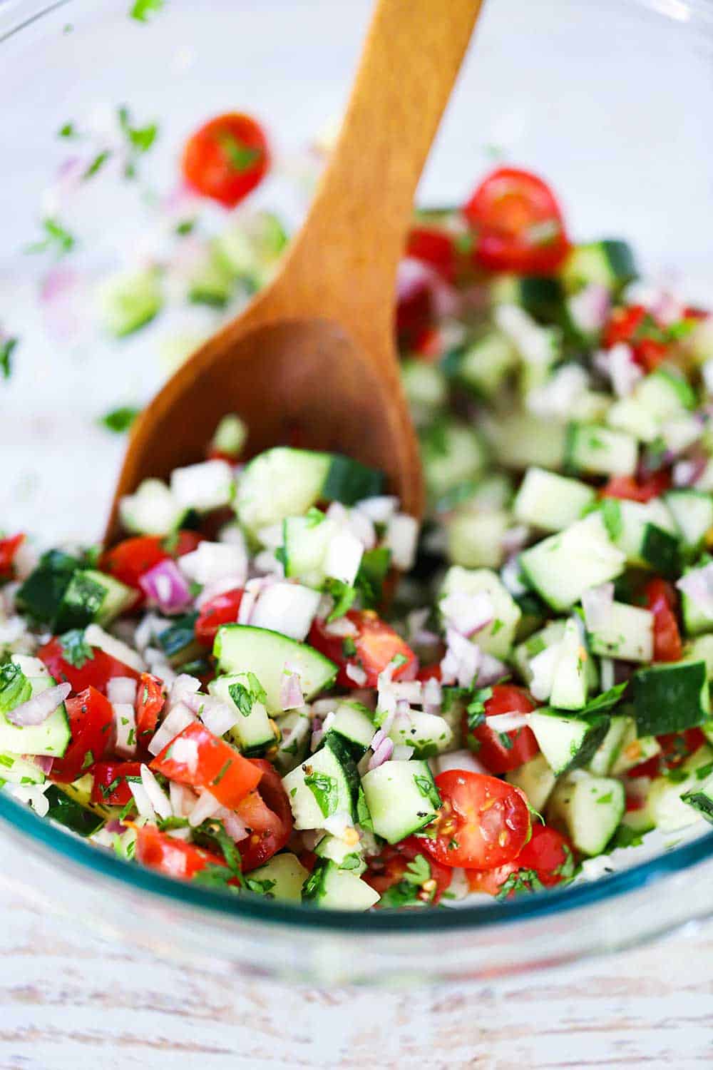 A large glass bowl filled with a cucumber and tomato salad with a large wooden spoon in it.