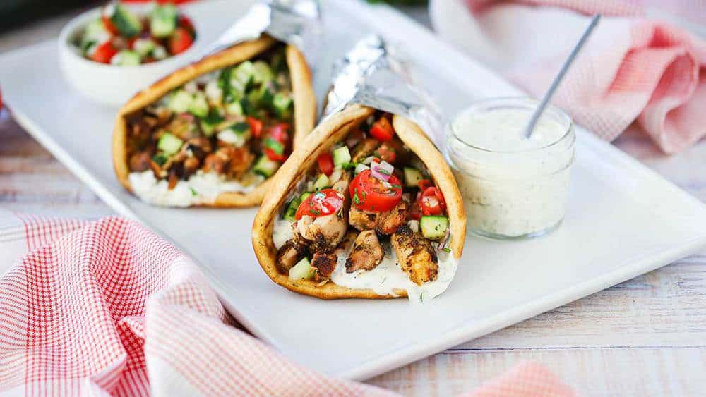 Two chicken gyros wrapped in foil on a white platter next to a small jar filled with tzatziki sauce with a spoon in, with checkered napkins nearby.