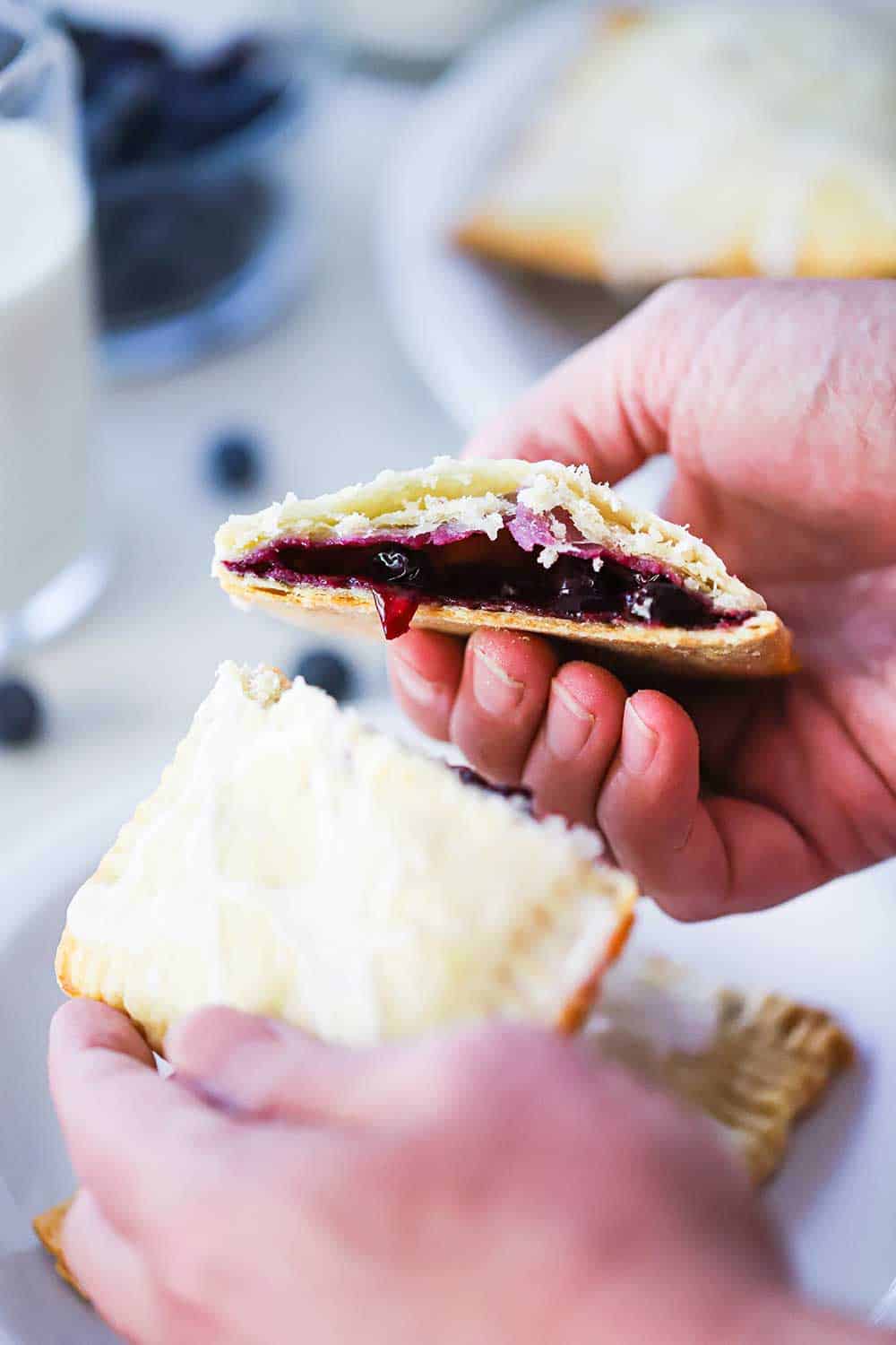 Two hands that are holding a blueberry pop tart that has been split in half.