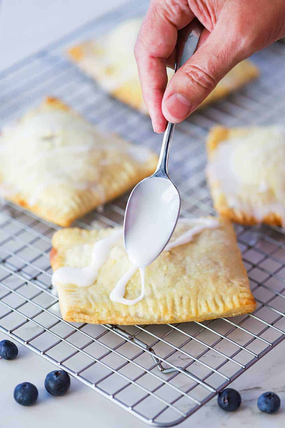 A hand using a spoon to drizzle sugar glaze over the top of baked homemade blueberry pop tarts.