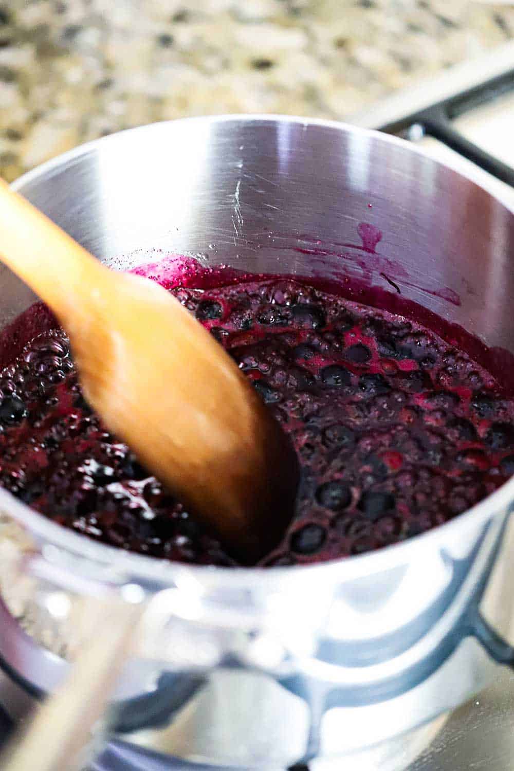 A silver saucepan filled with bubbling blueberry filling with a wooden spoon in the pan.