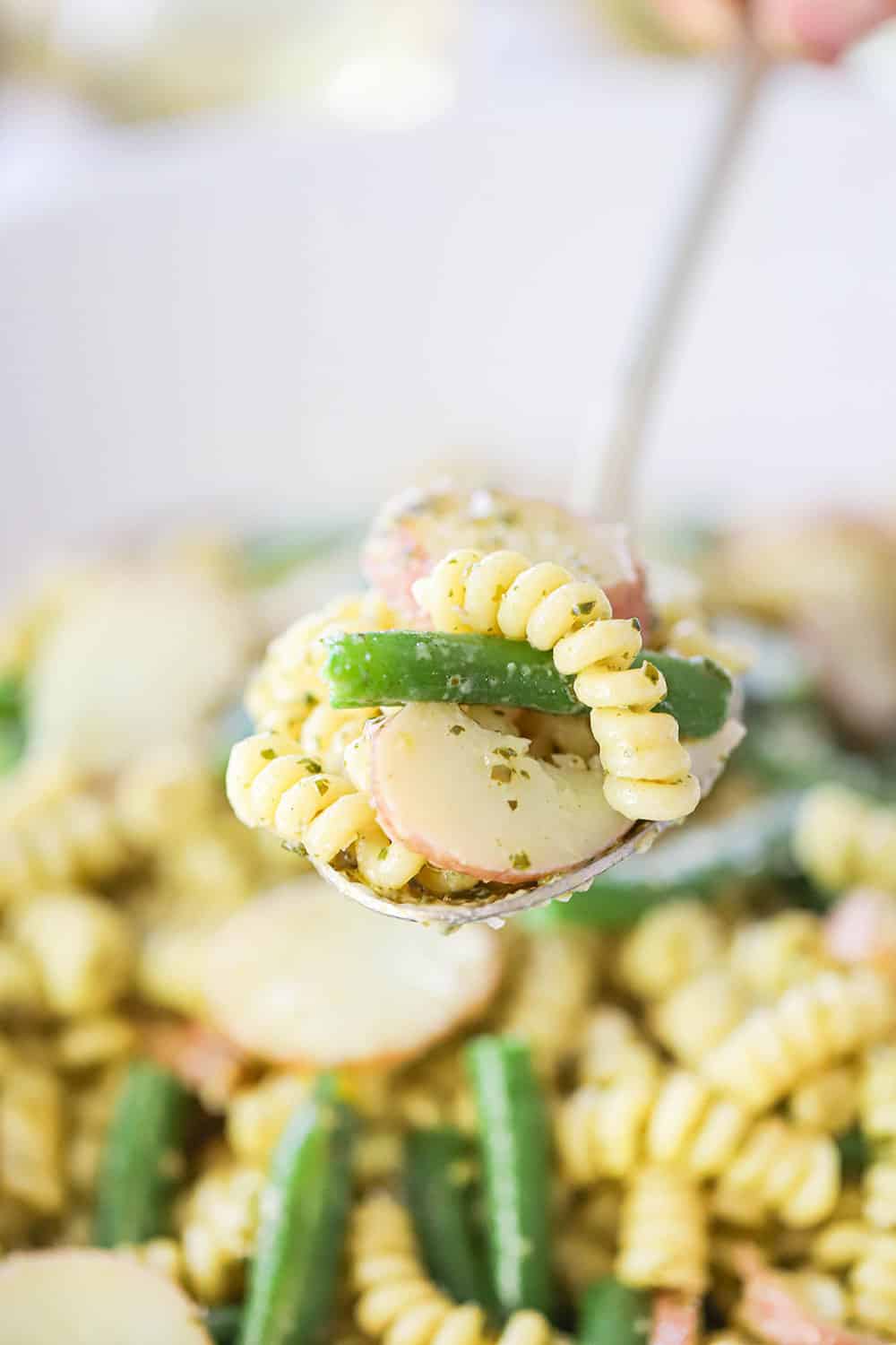 A hand holding up a large spoon filled with pesto pasta salad with potatoes and green beans.