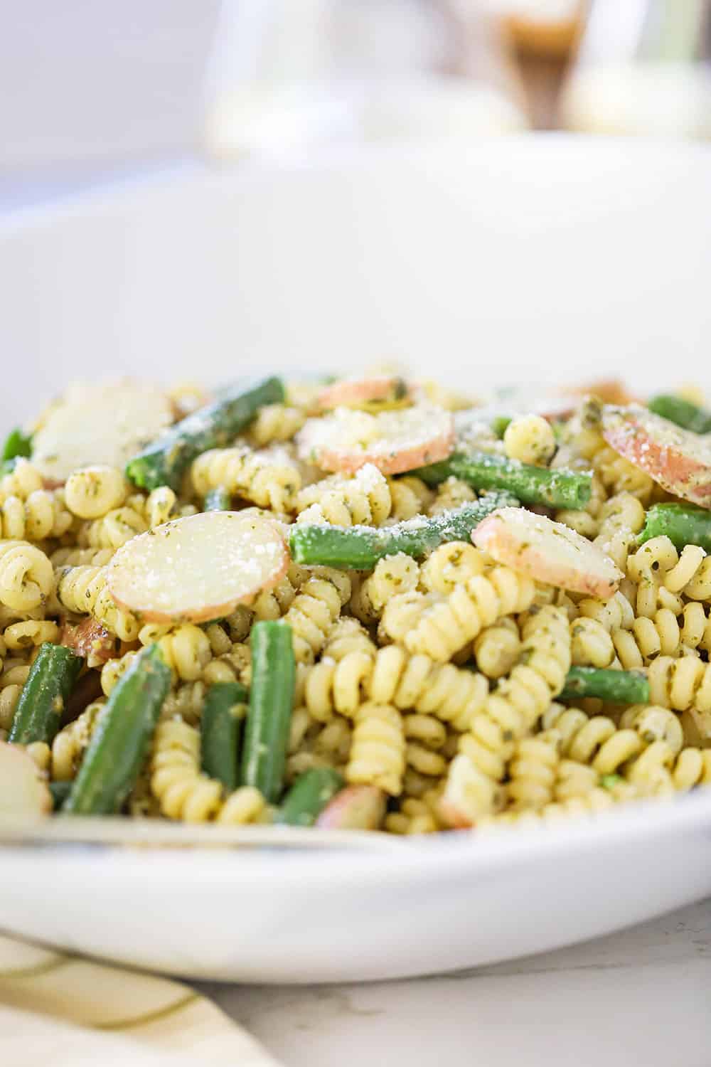 A close up view of a large circular white serving bowl filled with pesto pasta salad with sliced potatoes and green beans.