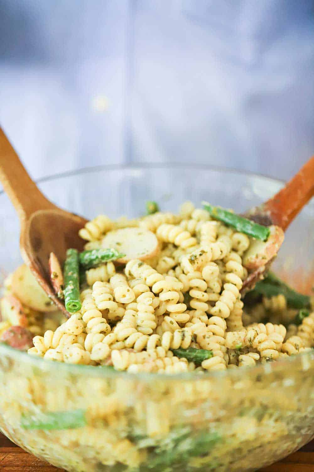 A person holding two wooden spoons in a glass bowl filled with pasta salad with green beans.