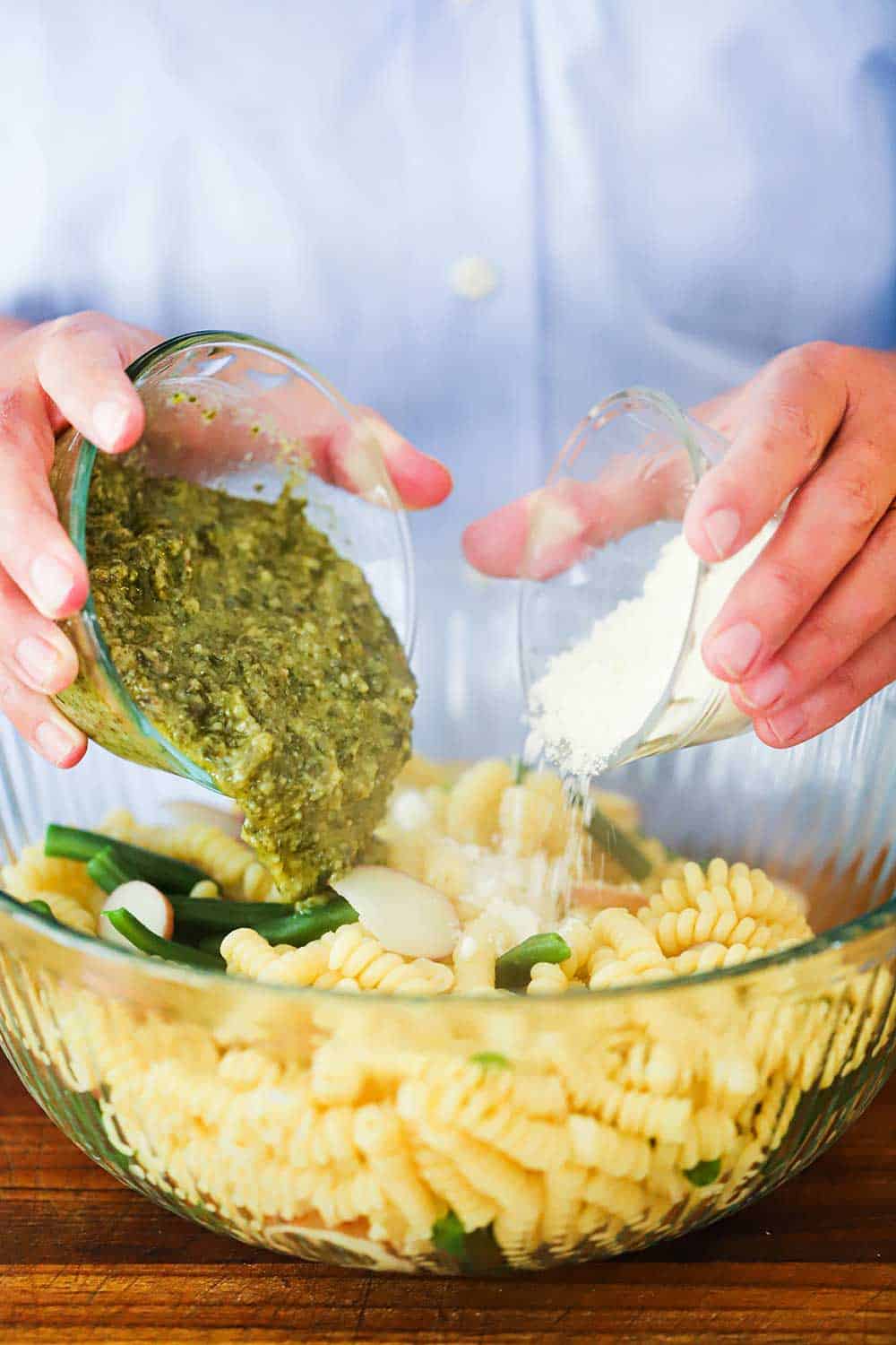 Two hands pouring a glass bowl of pesto sauce and another bowl of Parmesan cheese into a bowl of cooked fusili pasta.