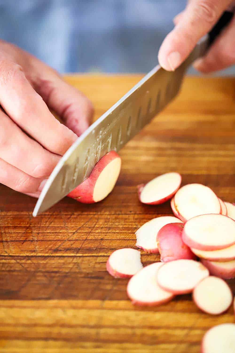 A hand holding a small red potato on a wooden cutting board while the other hand uses a knife to slice it, sitting next to a pile of sliced potatoes.