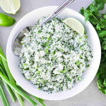 An overhead view of a large white bowl filled with Cilantro Lime Rice next to scallions and a bunch of cilantro, with a silver spoon in the bowl.