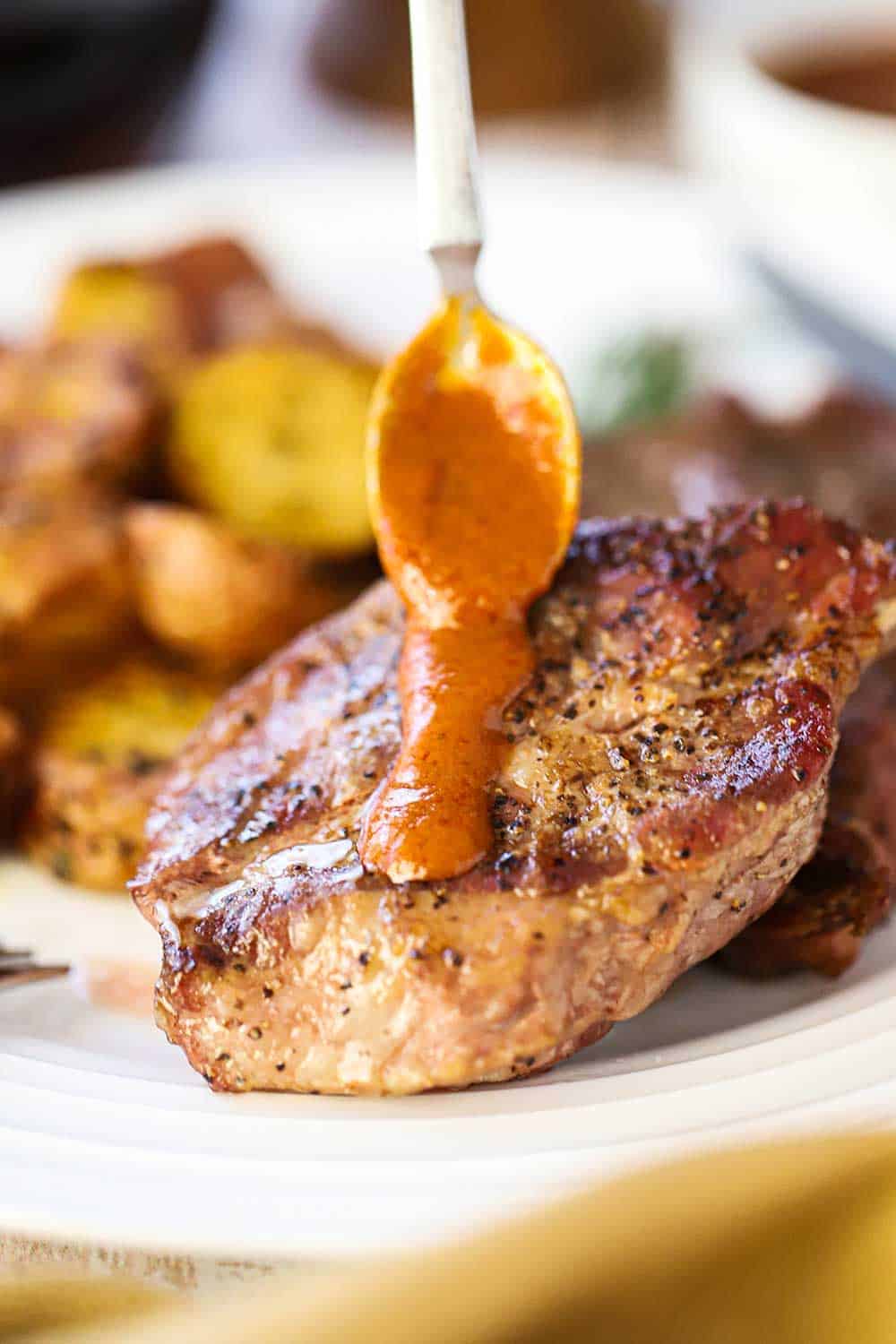 A small spoon drizzling homemade steak sauce down the top and center of a grilled steak on a white plate next to roasted potatoes. 