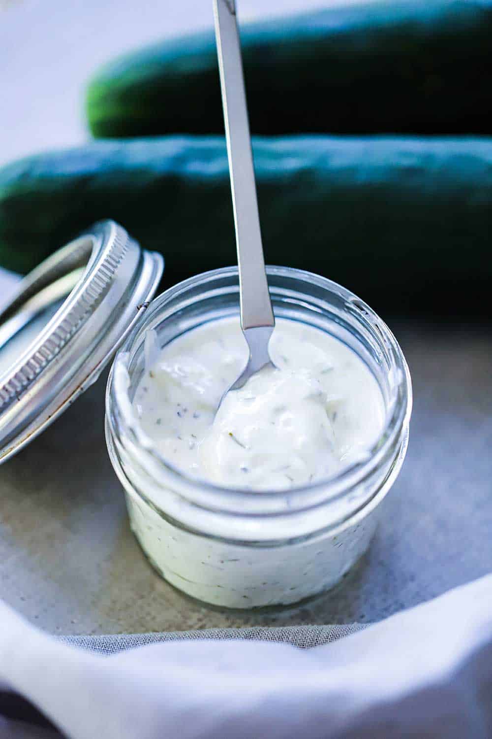 A small jar filled with homemade tzatziki sauce with a small spoon in it, next to two cucumbers and a grey napkin.