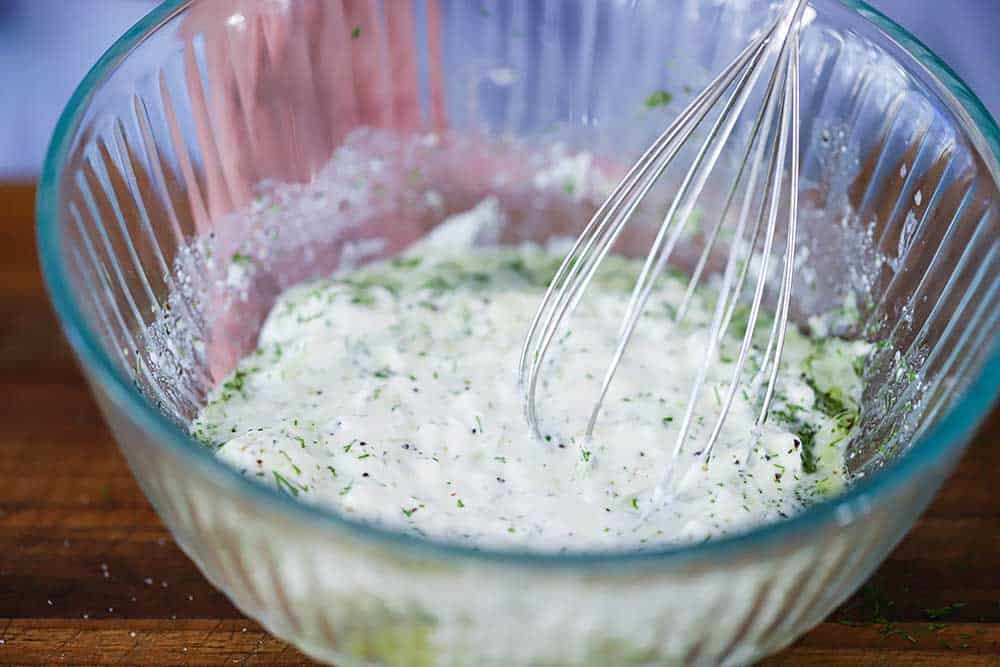A glass bowl filled withe the ingredients of homemade tzatziki sauce with a whisk in it.