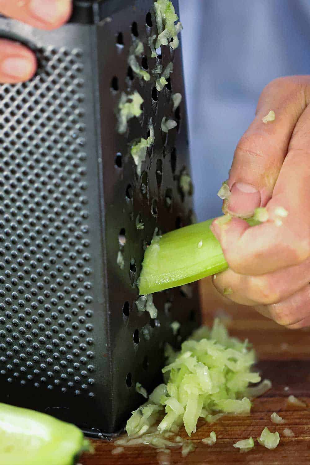 A hand scraping a peeled and seeded cucumber down the side of a box grater with shredded cucumber next to it on a cutting board.