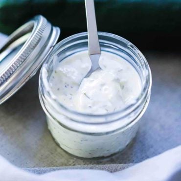 A small jar filled with homemade tzatziki sauce with a small spoon in it, next to two cucumbers and a grey napkin.