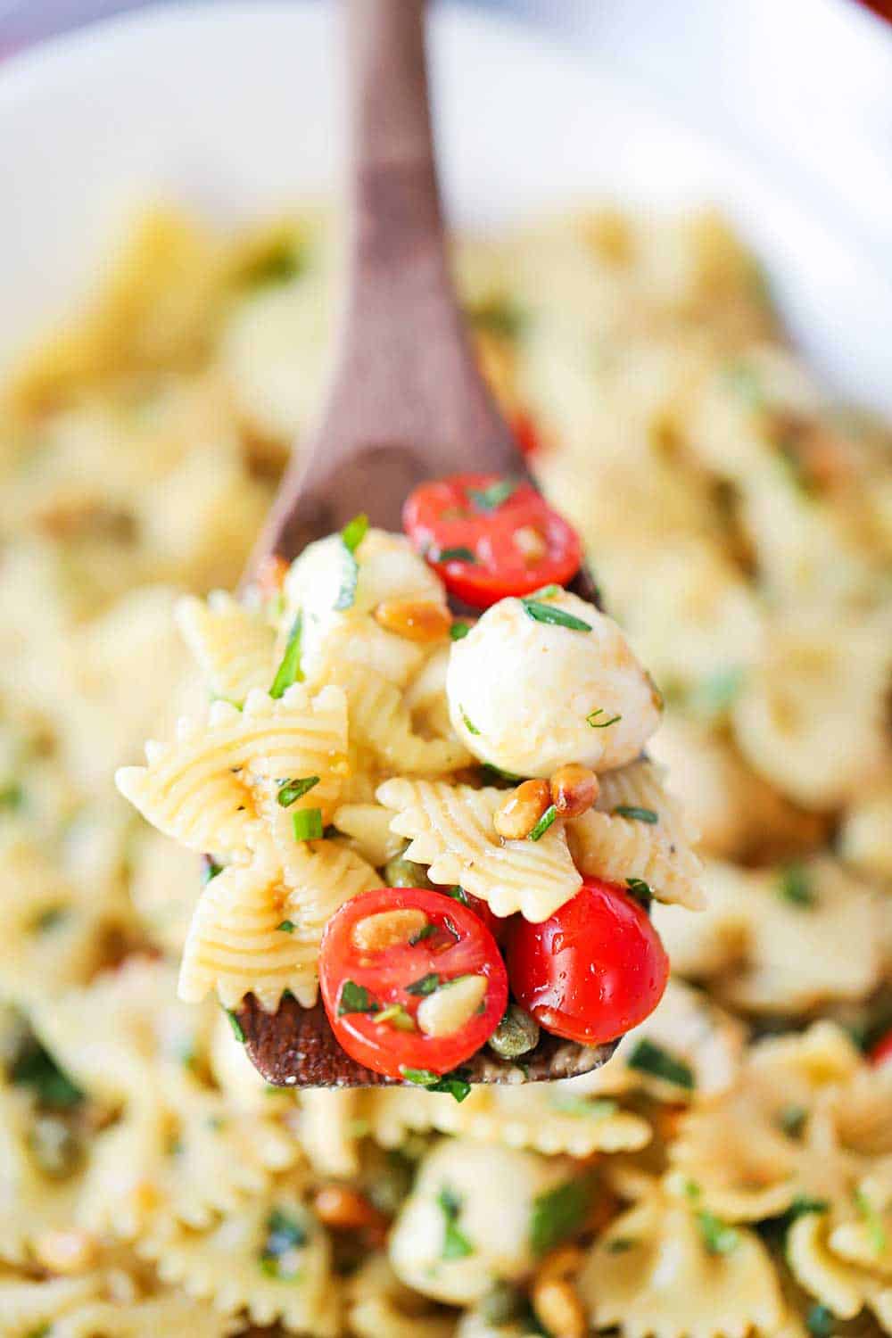 A large wooden spoon holding up a helping of Caprese pasta salad over a large bowl holding the salad.