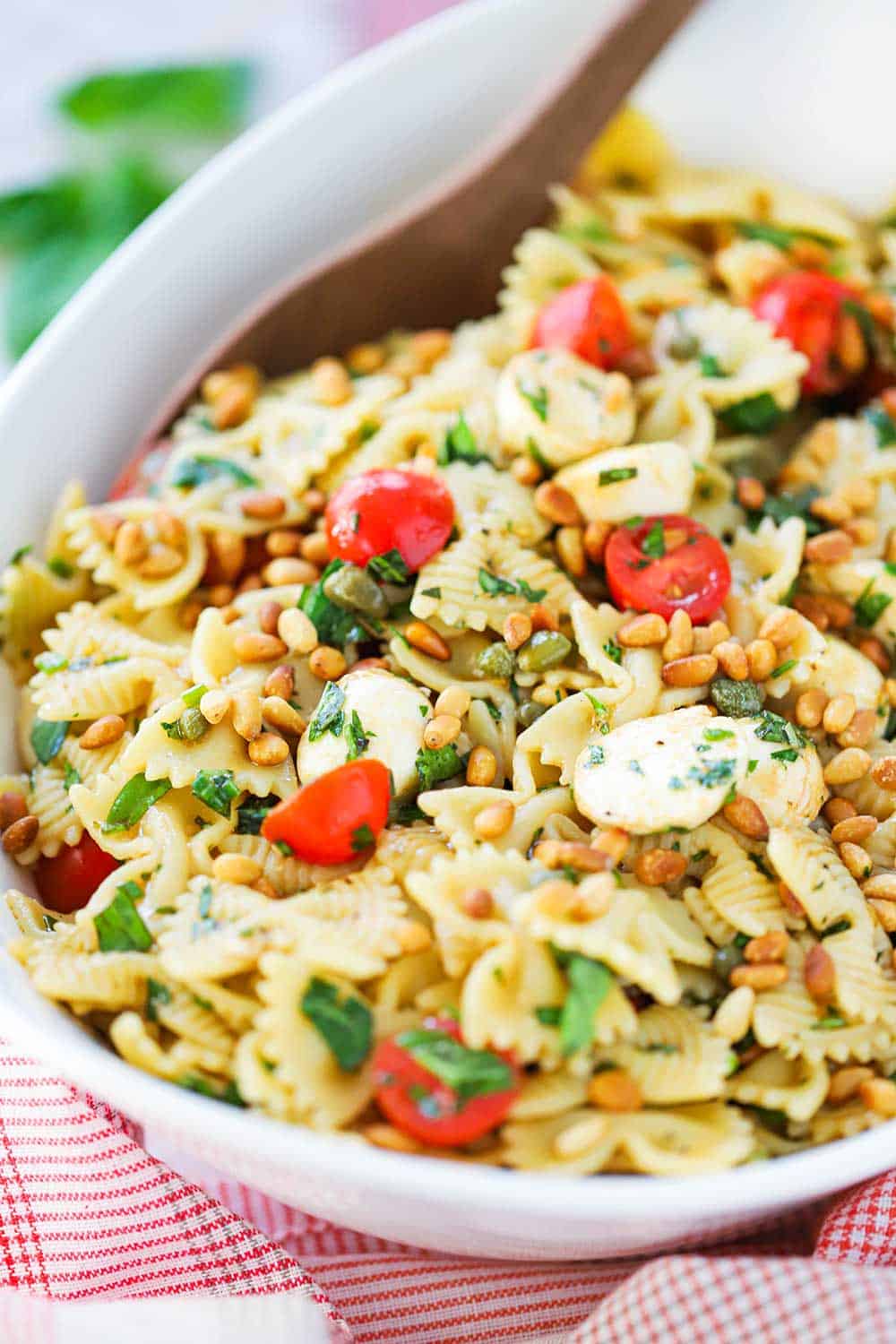 A close-up view of a caprese pasta salad in a large white serving bowl with a wooden spoon in the salad.