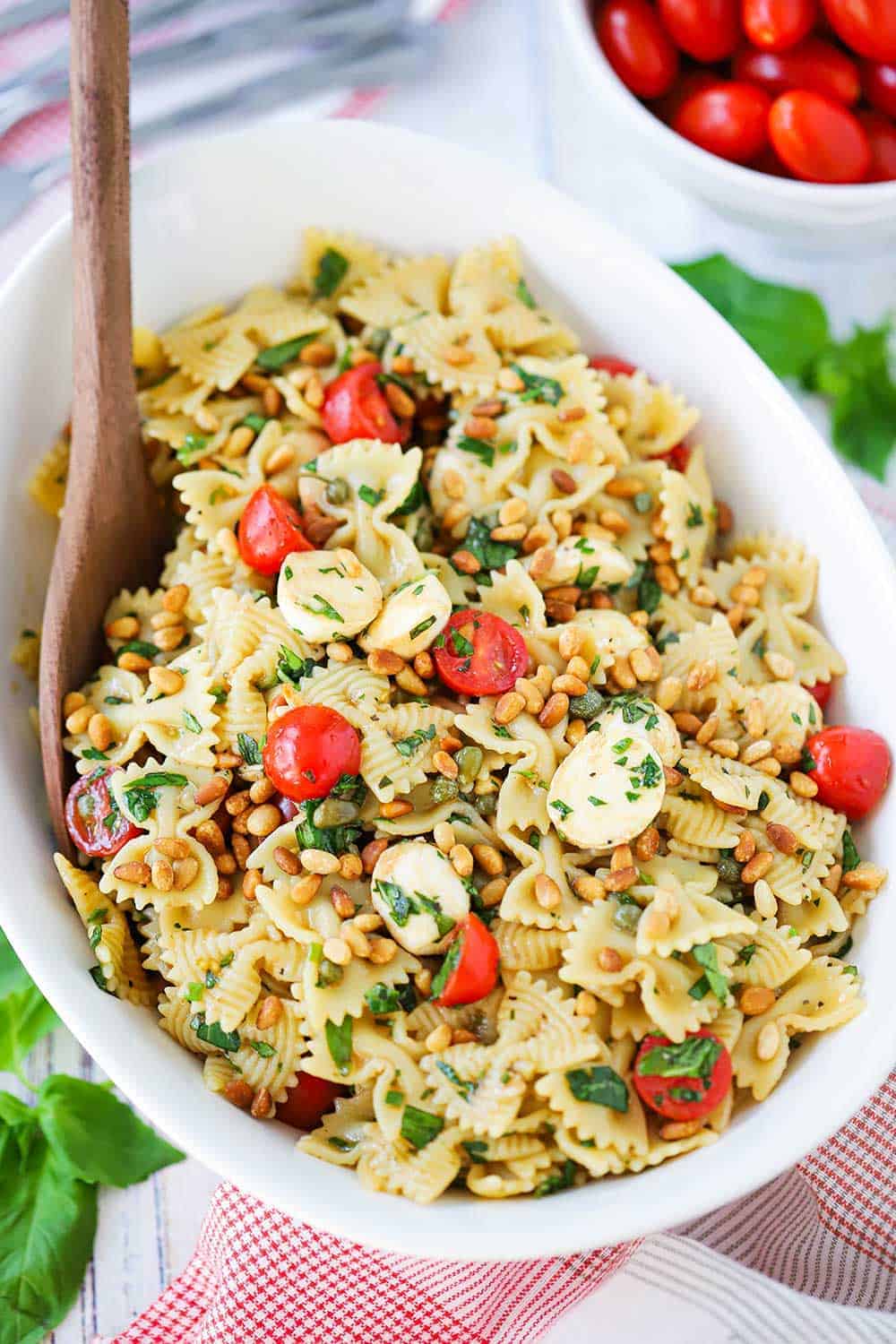 An overhead view of a white serving bowl filled with a Caprese pasta salad with a wooden spoon in the salad next to a patterned napkin and a bowl of cherry tomatoes.