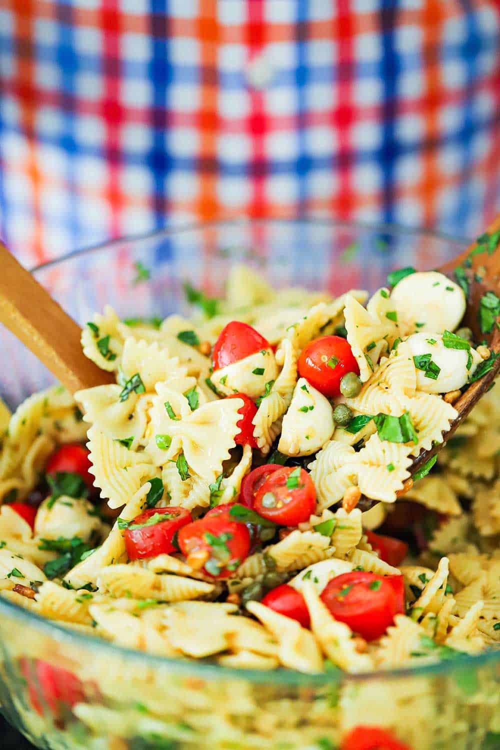 A person using two wooden spoons to mix together a caprese pasta salad in a large glass bowl.