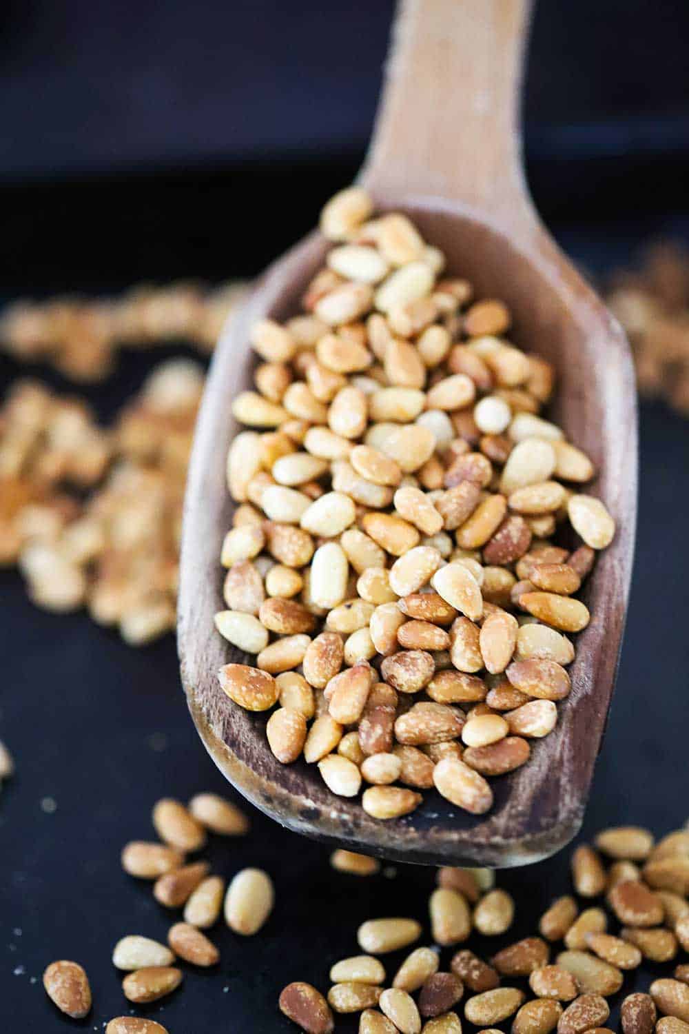 A large wooden spoon being held up and filled with toasted pine nuts over a pan with pine nuts in it.