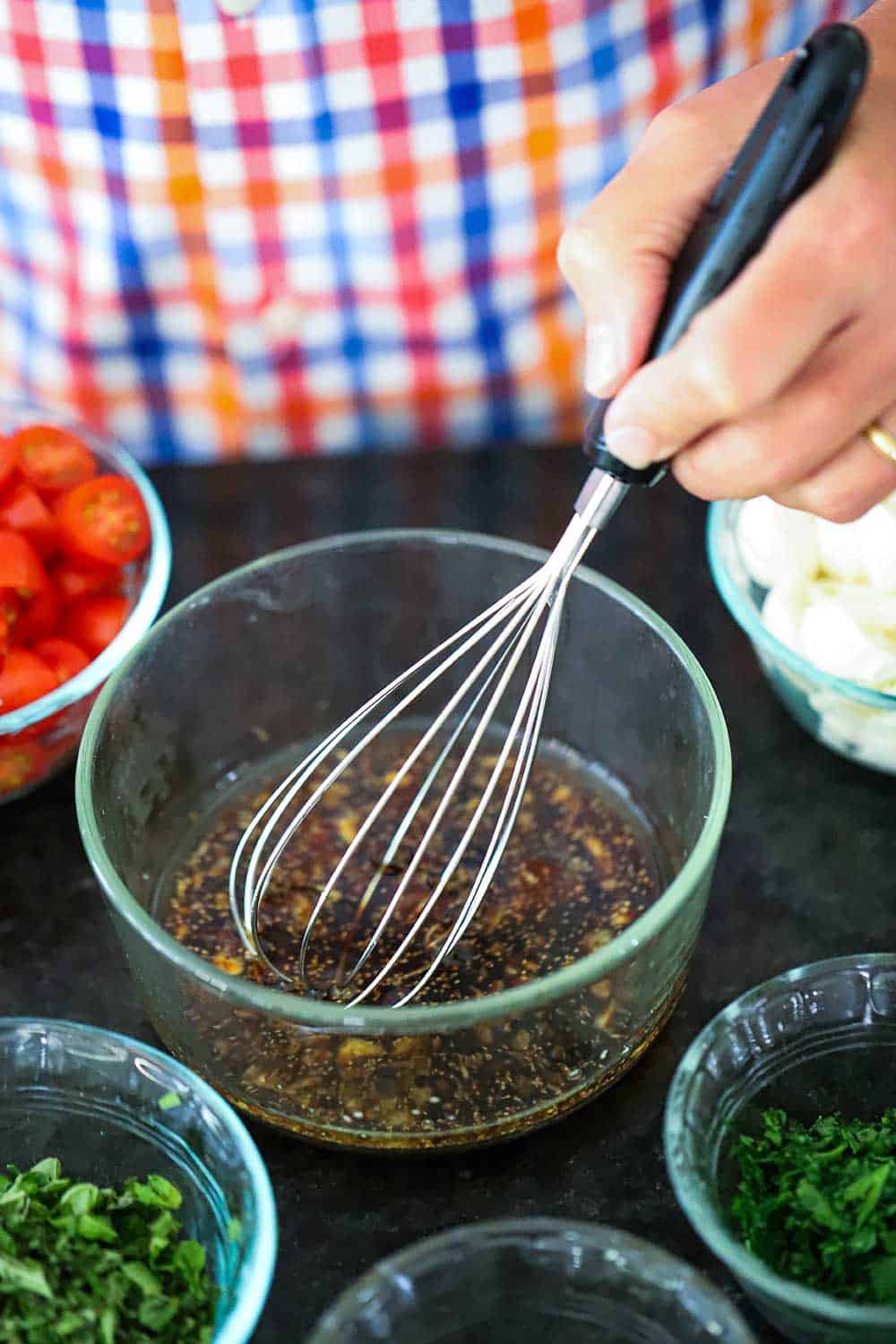 A hand using a whisk to combine a marinade in a glass bowl next to small glass bowls of herbs, mozzarella, and tomatoes.