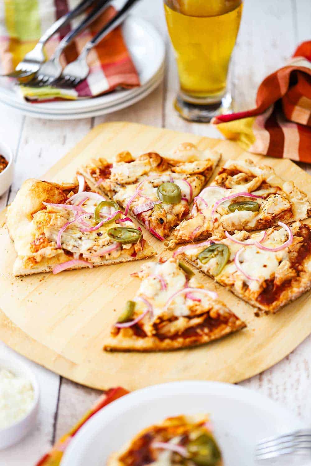 A side view of a BBQ chicken pizza cut into slices with a piece missing, and a glass mug of beer nearby and checkered napkins.