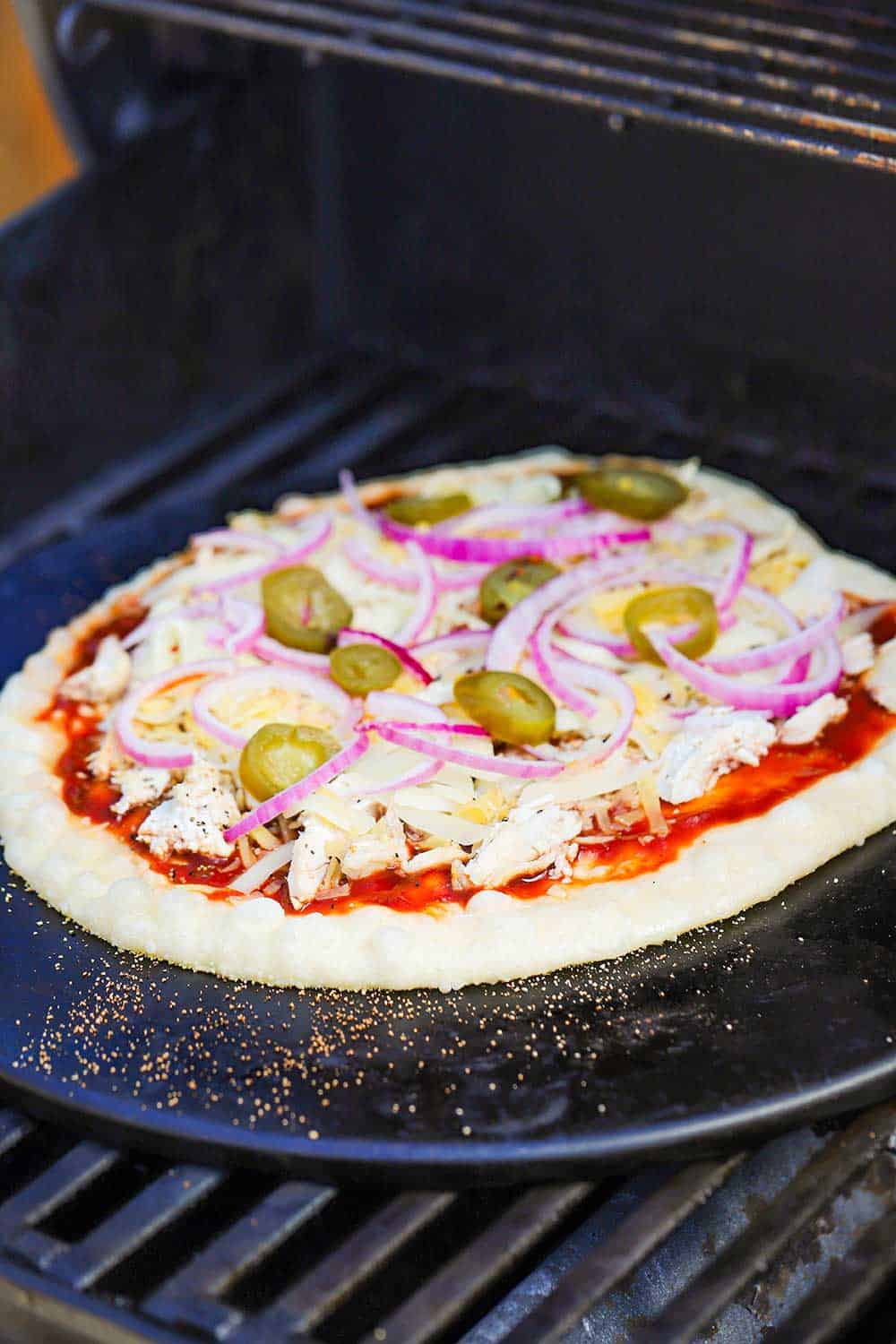 An uncooked pizza topped with BBQ sauce, shredded chicken, cheese, red onion, and jalapeno slices all on a pizza stone sitting on the grates of a gas grill.