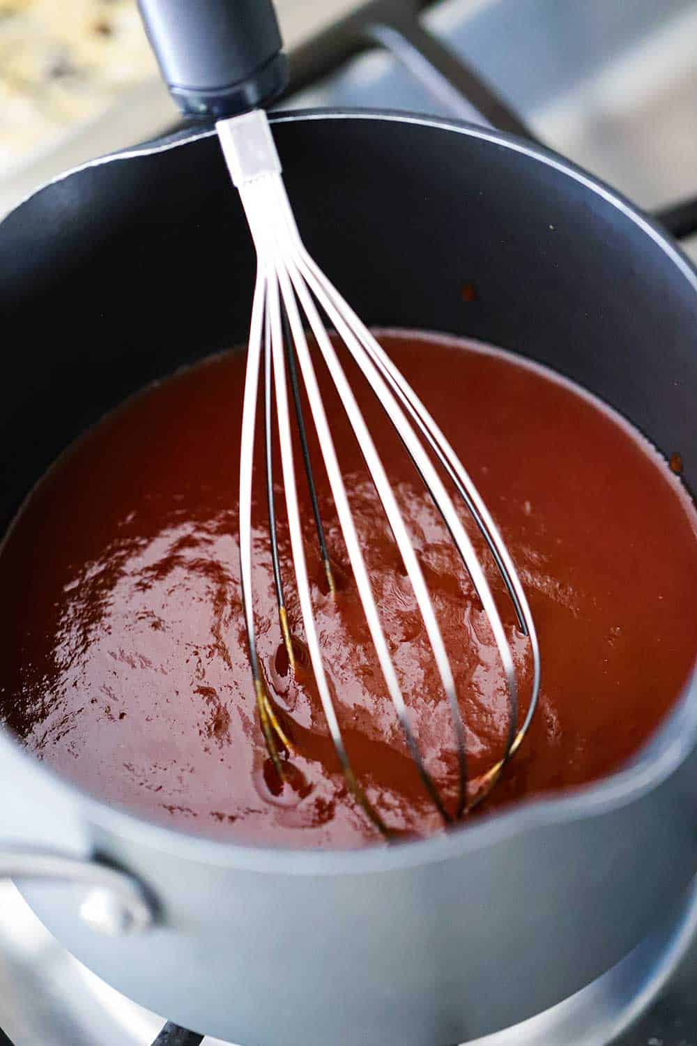 An overhead view looking into a medium saucepan filled with BBQ sauce with a whisk in it.