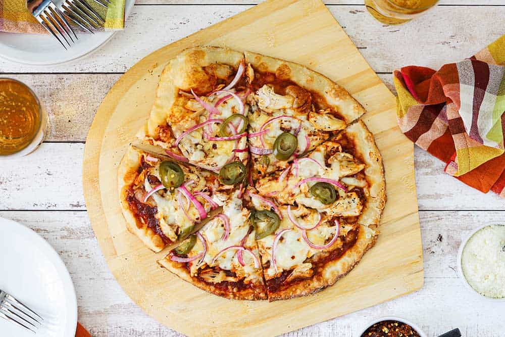 An overhead view of a sliced BBQ chicken pizza on a pizza paddle next to a white plate, a glass of beer, and a checkered napkin.