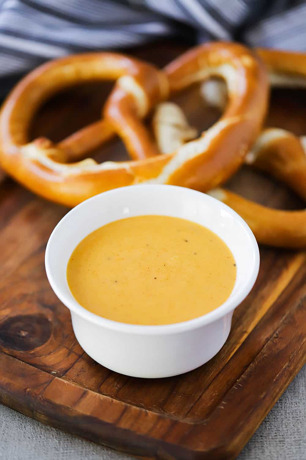 A small cutting board with two large pretzels in the background and a bowl of beer cheese sauce in the foreground. 