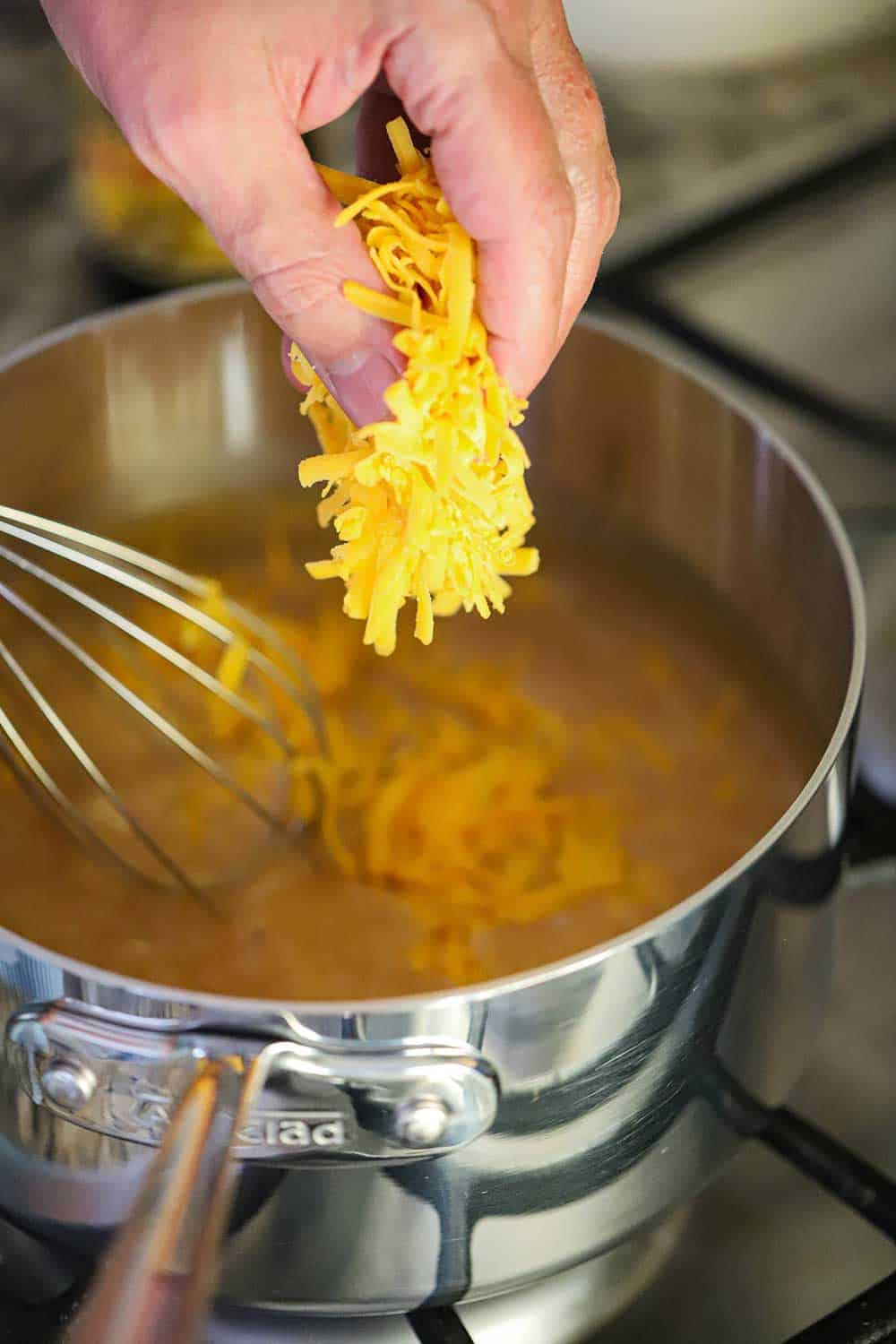 A hand dropping shredded cheddar cheese into a sauce pan with a beer sauce. 