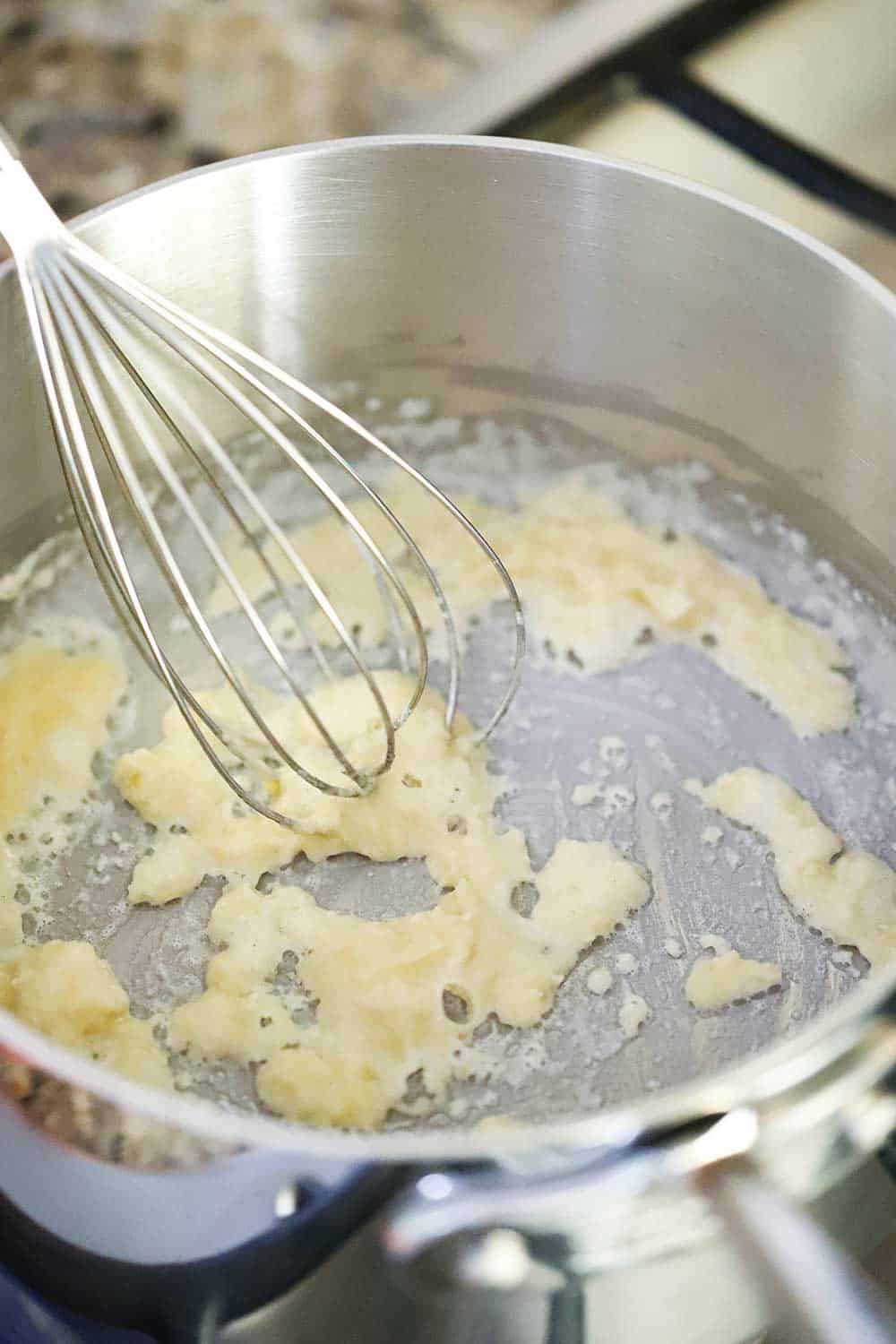 A whisk stirring a white roux in a stainless steel saucepan. 