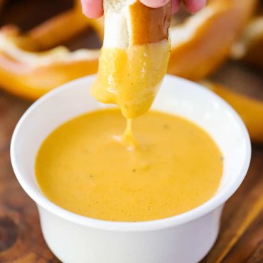 A hand dipping a piece of pretzel into a white bowl of beer cheese sauce.