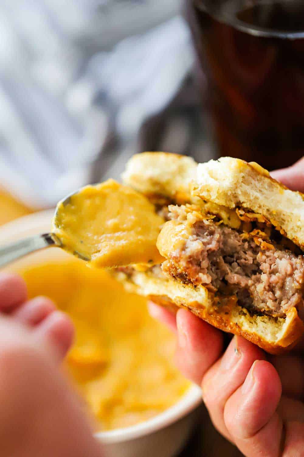 A hand holding a slider with another hand spooning cheese sauce on it. 