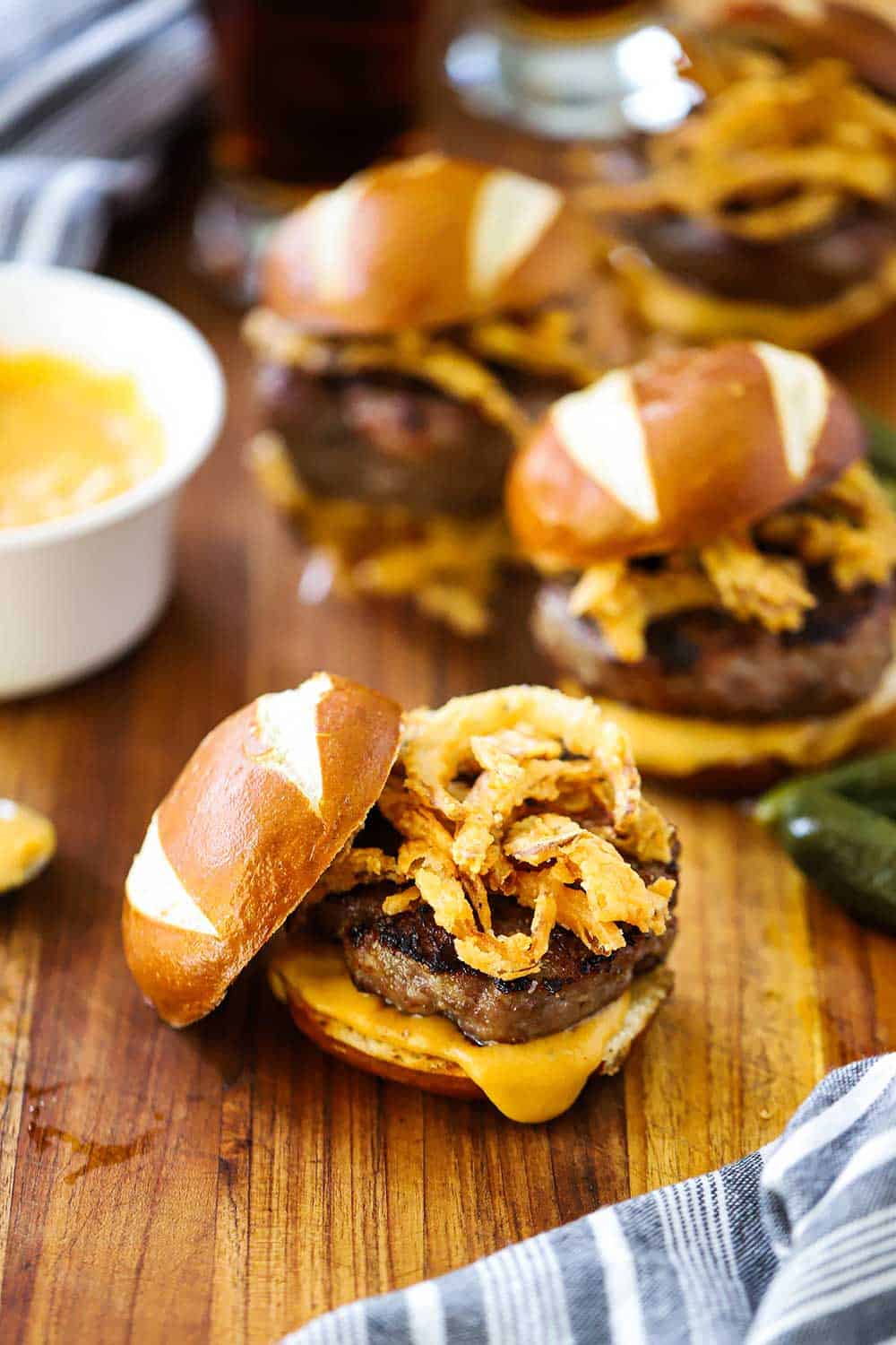 A cutting board with multiple bratwurst sliders on them with fried onions on top and cheese sauce nearby. 