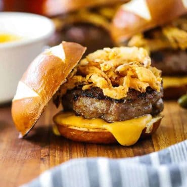 A bratwurst slider on a pretzel bun with cheese sauce and fried onions on a cutting board.