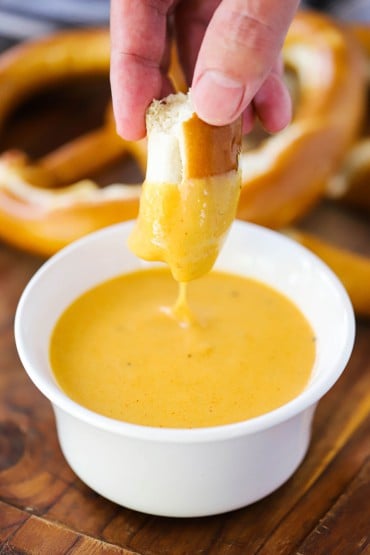A hand dipping a piece of pretzel into a white bowl of beer cheese sauce.
