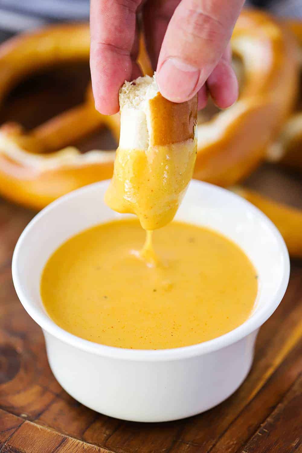 A hand dipping a piece of pretzel into a white bowl of beer cheese sauce. 