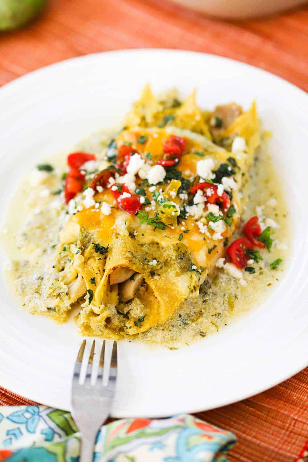 A white plate filled with two chicken enchiladas with verde sauce topped with chopped tomatoes, cilantro and Mexican cheese, with a fork on the side of the plate. 