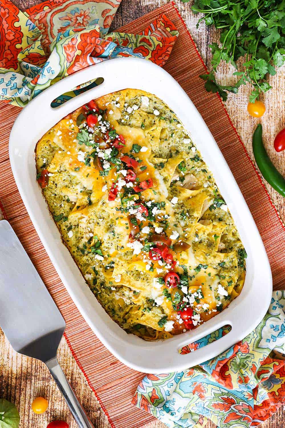 A 9'x13' white baking dish filled with chicken enchiladas with verde sauce topped with chopped tomatoes and cilantro, alongside a metal spatula and festive napkins. 