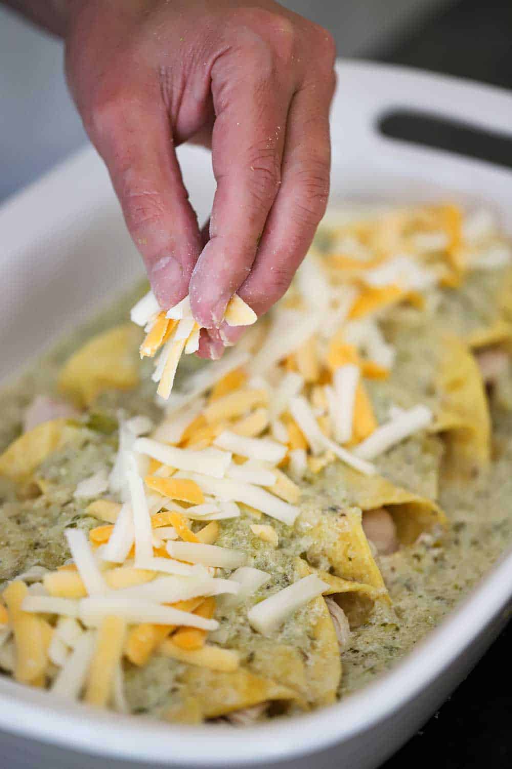 A hand sprinkling shredded Mexican cheese over the tops of chicken enchiladas with verde sauce in a white baking dish. 