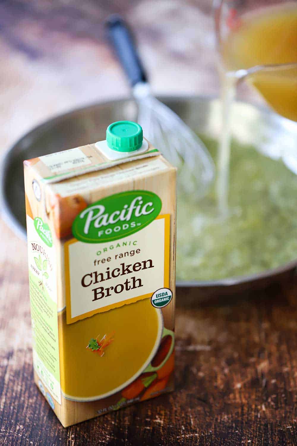 A box of Pacific Food Chicken Broth in front of a silver skillet filled with verde sauce with a whisk in it and a measuring cup pouring in the broth. 