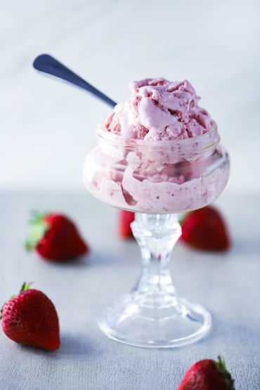 A glass ice cream holder filled with homemade strawberry ice cream with a spoon in it and whole strawberries scattered around next to it.