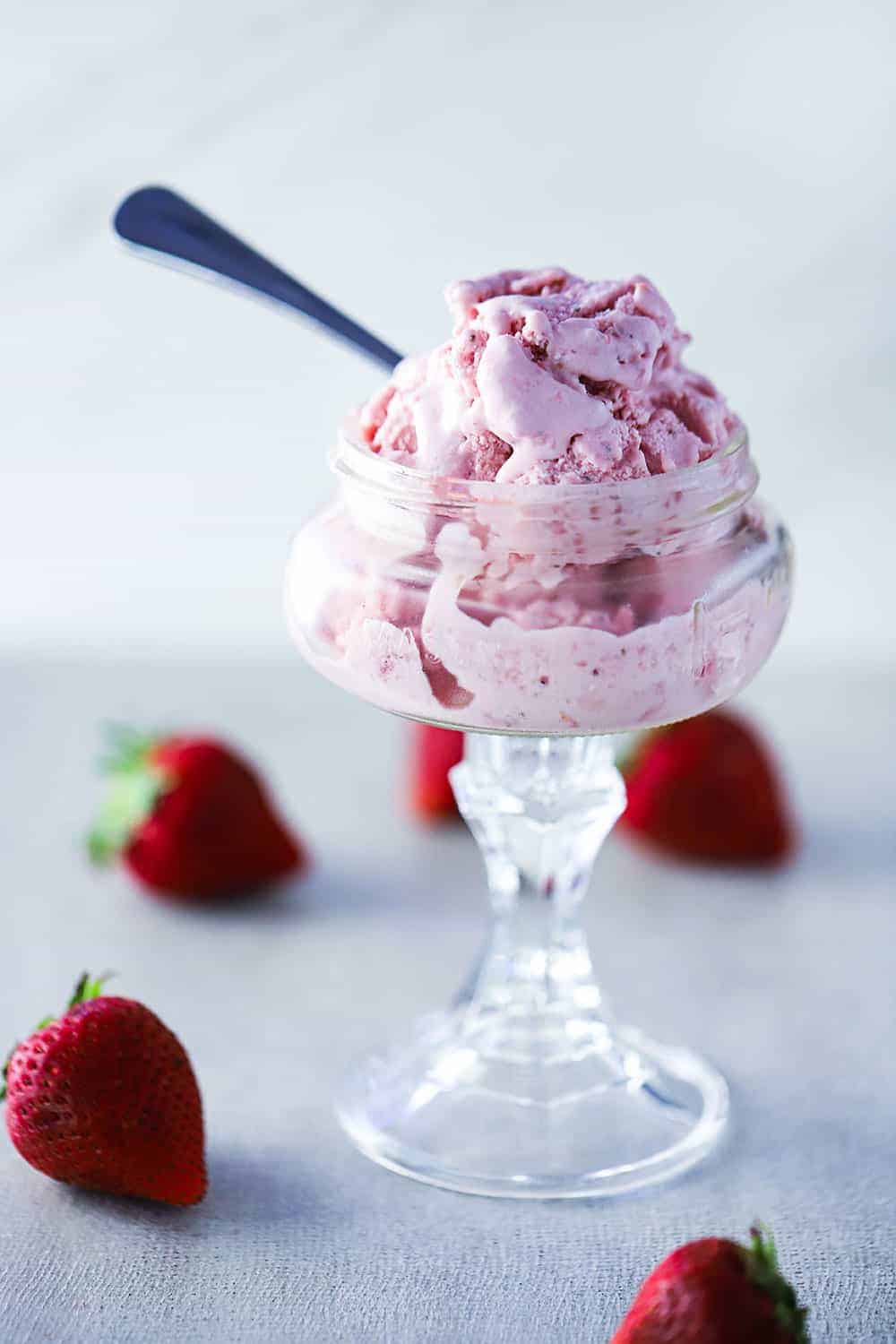 A glass ice cream holder filled with homemade strawberry ice cream with a spoon in it and whole strawberries scattered around next to it. 