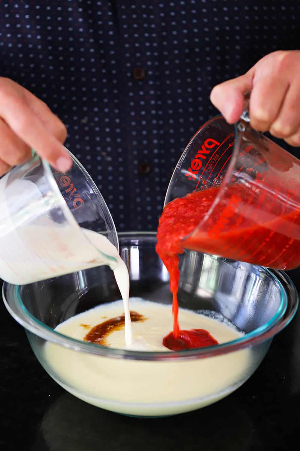 A person using both hands to pour pureed strawberries and heavy cream into a bowl of custard. 