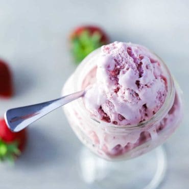 An overhead view of a glass bowl with a stem holding a serving of homemade strawberry ice cream with whole strawberries scattered around next to it.