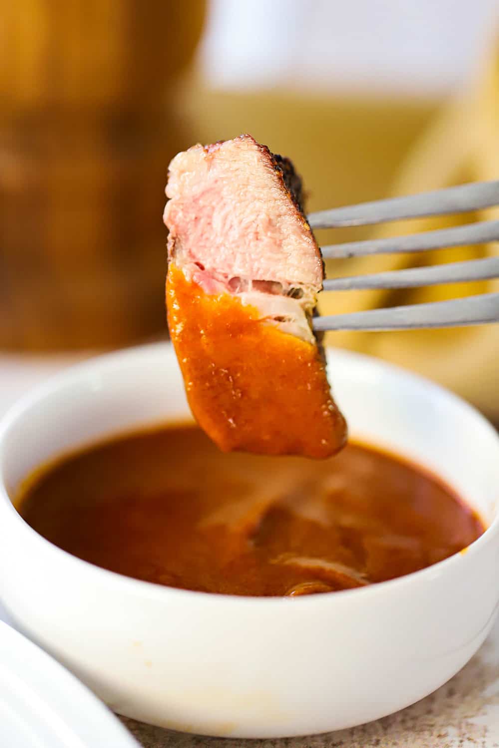 A fork with a slice of pork steak stuck into that is over a small white bowl of steak sauce that it has been dipped into.